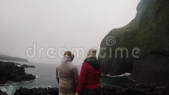 雾天年轻游客夫妇穿着雨衣站在海边欣赏风景视频的预览图