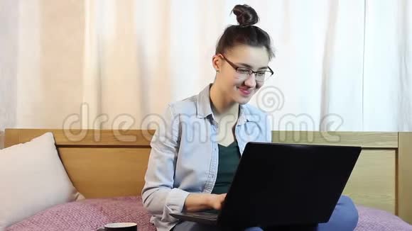 一个年轻迷人的女孩正在从她的笔记本电脑发短信坐在家里的床上视频的预览图