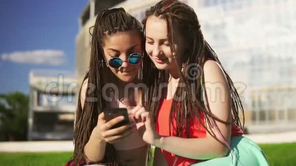 快乐的女人在夏天的公园里坐在草地上用智能手机年轻的朋友在聊天和看视频的预览图