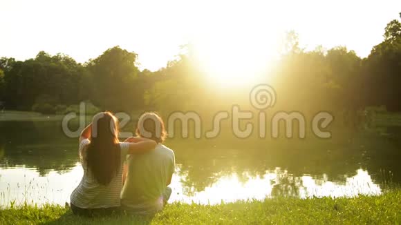 可爱的女孩和男孩坐在湖边的绿草上享受日落回到户外幸福夫妇的视野视频的预览图