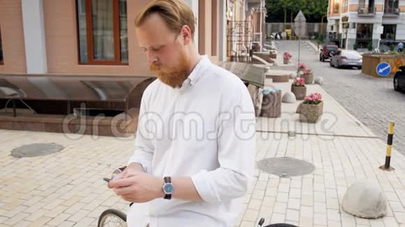 4k镜头穿着白色衬衫的时髦红胡子男子坐在老式自行车上并在手机上输入信息视频的预览图