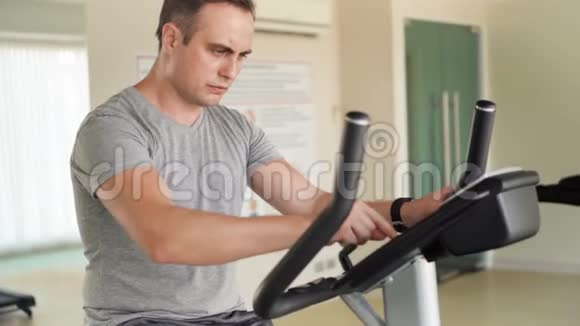 坚定帅气的年轻人穿着灰色t恤在健身房骑着健身车做运动视频的预览图