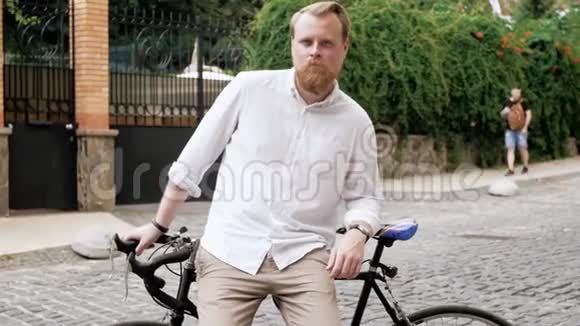 4K镜头穿着白色衬衫骑自行车在老街上的时髦留胡子男子视频的预览图
