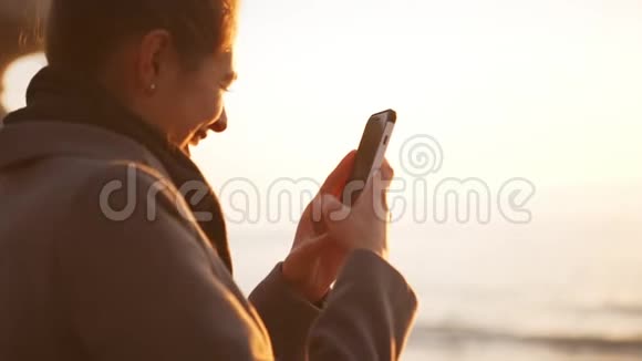 快乐的年轻女士用智能手机慢镜头拍摄海上日出太阳耀斑视频的预览图