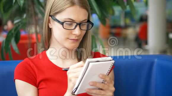 聪明的微笑学生穿着红色T恤和眼镜坐在咖啡馆里在笔记本上写字视频的预览图