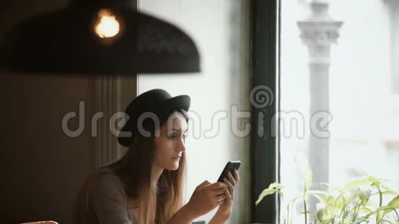 时尚的年轻女性戴着帽子在咖啡馆里使用智能手机有魅力的女性坐在窗边上网视频的预览图