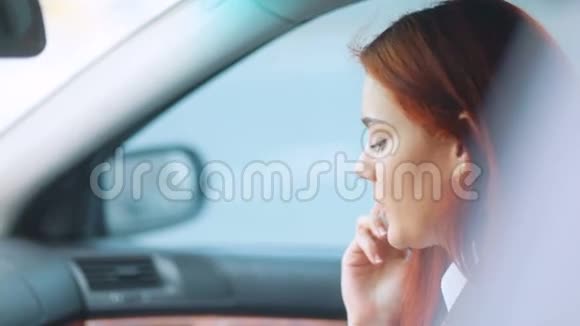 穿着浅棕色夹克和白色t恤的红发女孩坐在车里在智能手机旁讲话真是太棒了视频的预览图