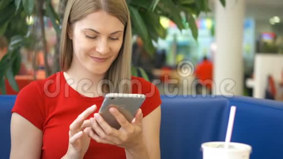 穿着红色T恤的年轻女子坐在咖啡馆里用智能手机与朋友聊天的画像视频的预览图