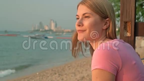 年轻漂亮的梦幻女郎穿着粉红色的t恤在海滩海上日落船城市景观背景视频的预览图