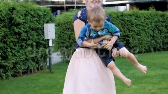 欢快的年轻妈妈在公园里抱着蹒跚学步的儿子在草地上旋转的慢动作视频视频的预览图