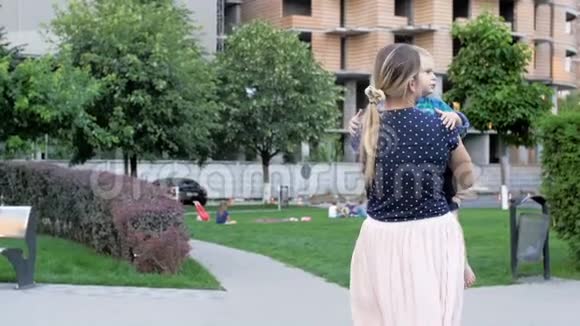 美丽的年轻女子抱着蹒跚学步的儿子在公园散步的慢镜头视频的预览图
