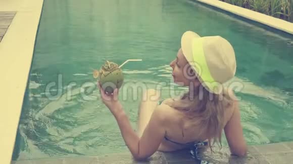 比基尼的年轻苗条的女人喝着热带度假胜地泳池边的可可椰奶复古视频的预览图