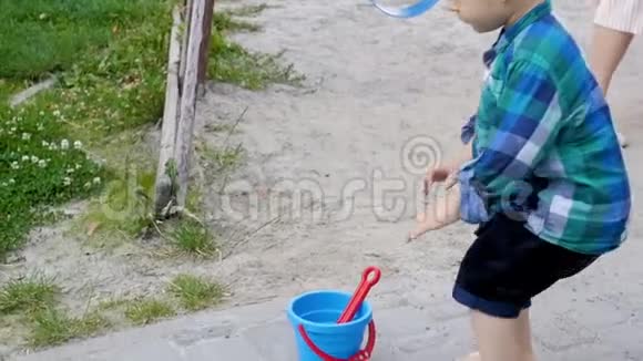 公园里赤脚学步的男孩在沙箱里拿着塑料桶和铲子的慢动作镜头视频的预览图