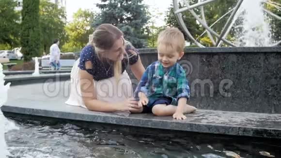 快乐的母亲带着蹒跚学步的儿子坐在喷泉旁泼水的慢镜头视频的预览图