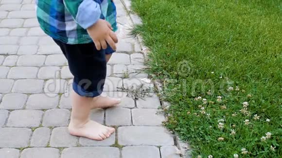光着脚的小男孩站在铺好的路上试图踩在公园的新鲜青草上的慢镜头视频的预览图