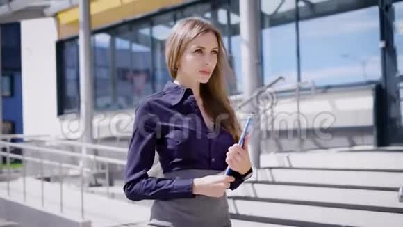 年轻漂亮的女士穿着蓝色衬衫和灰色裙子手里拿着剪贴板走在办公楼附近的户外视频的预览图