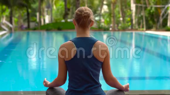 在热带的游泳池附近年轻女子坐在瑜伽垫上以莲花的姿势练习瑜伽视频的预览图