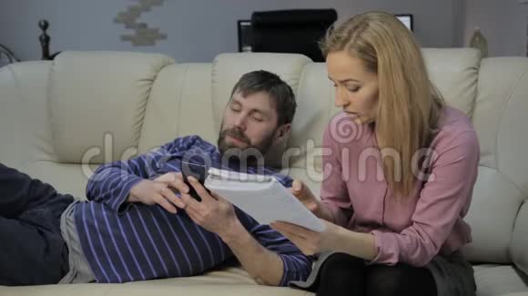 金发学生读她的笔记她的男朋友坐在沙发上用智能手机视频的预览图