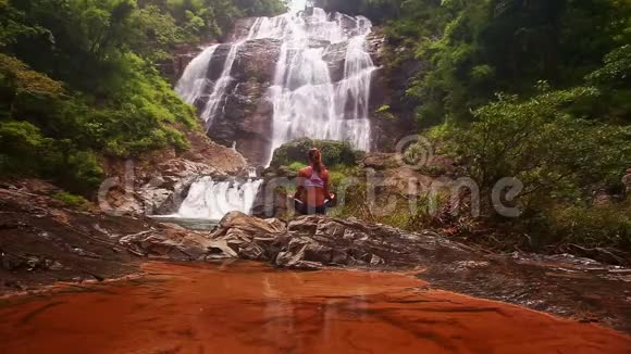 女孩坐在瀑布红色透明湖附近的瑜伽姿势视频的预览图
