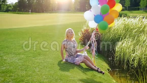 美丽的金发女孩带着五颜六色的气球坐在草地上的慢镜头视频的预览图