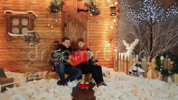 一对相爱的夫妇把一份红色的礼物扔到一座木制童话房子的门廊上圣诞节和新年主题视频的预览图