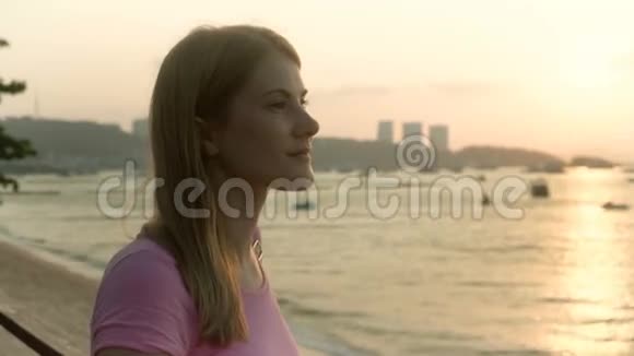 年轻漂亮的梦幻女郎穿着粉红色的t恤在海滩海上日落船城市景观背景视频的预览图