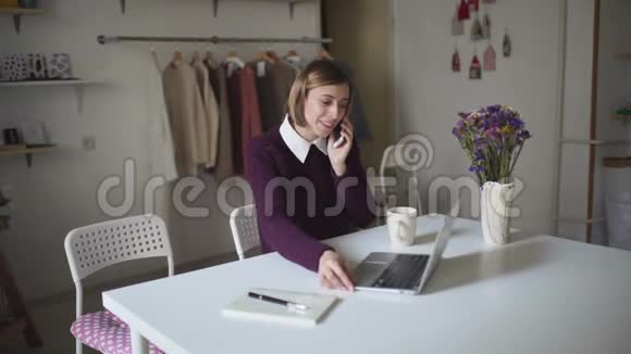 年轻女子坐在桌子旁用手机说话女人用手提电脑视频的预览图