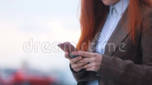 穿着棕色夹克和白色t恤的红发女孩的特写镜头轻敲她的智能手机在城市背景上微笑视频的预览图