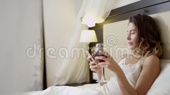 女人在床上喝白葡萄酒时在智能手机上发短信视频的预览图