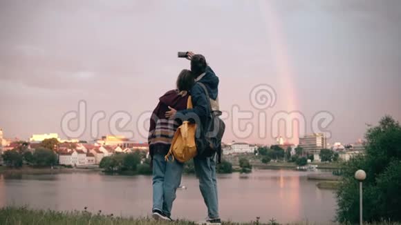 年轻漂亮的夫妇背着背包在智能手机上的铆钉岸边拍摄彩虹的照片视频的预览图