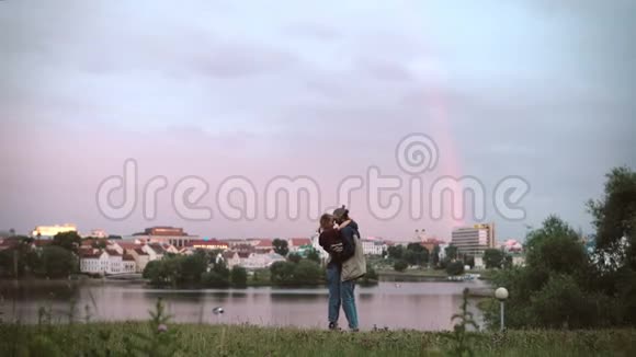 美丽的年轻男女站在河岸上拥抱在夕阳下彩虹在背景上视频的预览图