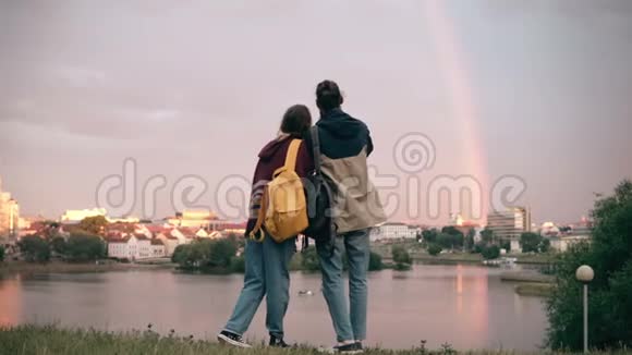 年轻的时髦夫妇拥抱在河岸上在智能手机上拍摄彩虹的背面视频的预览图