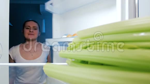 一个年轻漂亮的女人晚上从冰箱里拿和咬芹菜的4K镜头视频的预览图