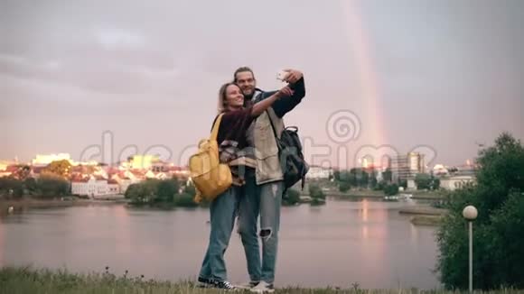 年轻的幸福夫妇拍摄的自拍照片与彩虹近河的背景微笑的男人和女人使用智能手机视频的预览图