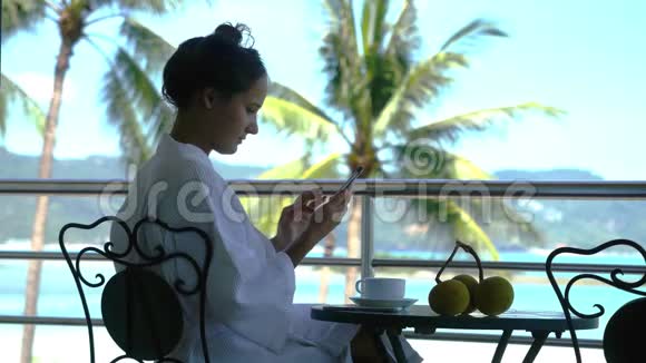 年轻的美女穿着浴袍坐在阳台上喝咖啡在令人惊叹的海景上使用手机视频的预览图