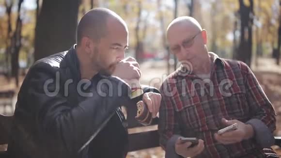 父亲和成年儿子坐在秋天公园的长凳上聊天与年迈的父亲在一个视频的预览图