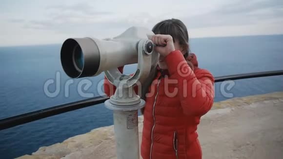 年轻的白种人女人穿着红色的泡泡夹克从高高的高架上望着海岸进入望远镜视频的预览图