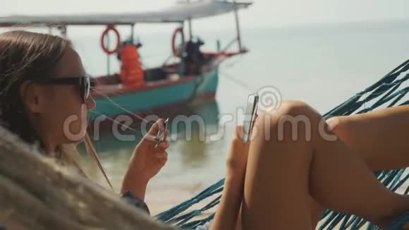 年轻女子在沙滩上用智能手机和银行卡躺在吊床上视频的预览图