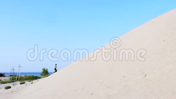 运动女孩穿着黑色的上衣和紧身裤跑上沙滩上升到沙地在荒芜的海滩上视频的预览图