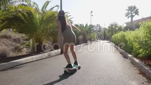 美丽的年轻女孩骑在靠近海滩的道路上棕榈树在长板上缓慢地运动视频的预览图