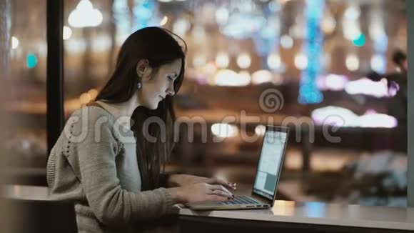 年轻漂亮的女人晚上坐在窗户附近用笔记本电脑工作女孩上网视频的预览图