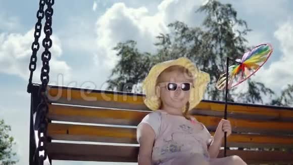 一个戴太阳镜的小可爱女孩在阳光明媚的温暖的日子里在跷跷板上摇摆视频的预览图