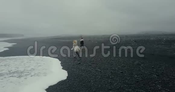 在冰岛黑色火山海滩散步的年轻夫妇的鸟瞰图洛帕皮萨的男人和女人把石头扔到海里视频的预览图