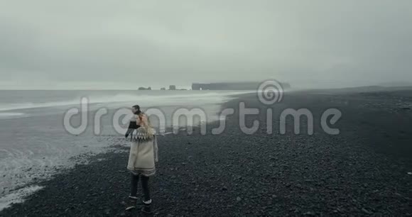 在冰岛黑色火山海滩散步的年轻夫妇的鸟瞰图洛帕皮萨的女人跳上男人的手臂视频的预览图