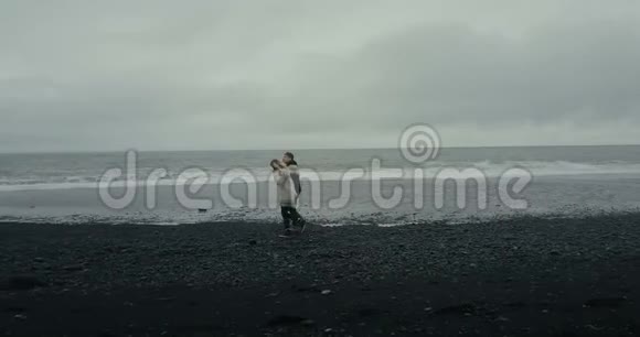 科普特乘坐冰岛毛衣在这对年轻夫妇附近飞行男人和女人走着双手握在黑色的火山海滩上视频的预览图