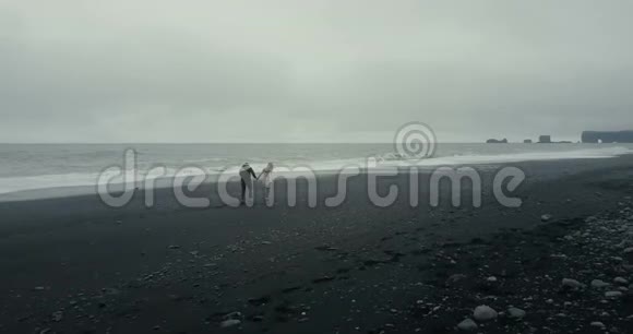 在冰岛黑色火山海滩上漫步的年轻夫妇的空中景色洛帕皮萨的游客享受海浪视频的预览图