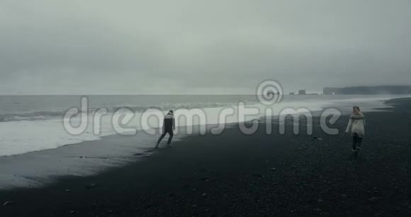 在冰岛的黑色火山海滩上向年轻夫妇鸟瞰男人从岸边的波浪中奔跑女人等待视频的预览图