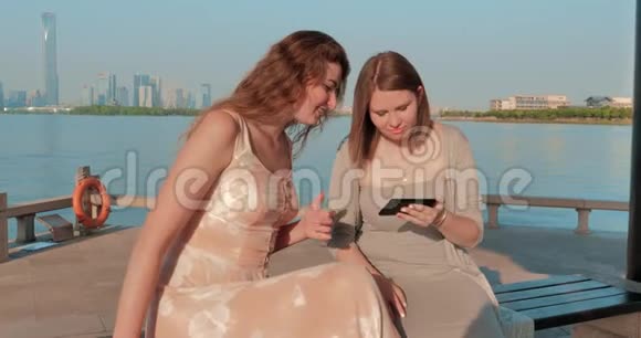 两个穿着浅色连衣裙的微笑女孩正在智能手机上看照片背景为蓝水湖和城市景观视频的预览图