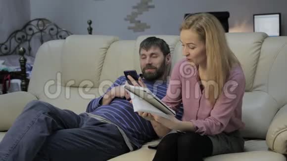 沙发上的金发妻子在笔记本上读笔记丈夫坐在旁边打电话视频的预览图