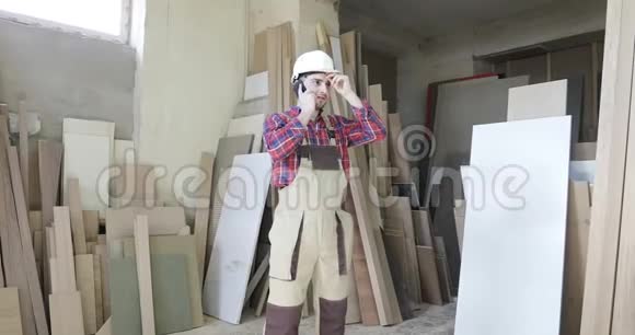一个穿细木工服的时髦木匠和一个白色头盔在家具厂的手机上交谈视频的预览图